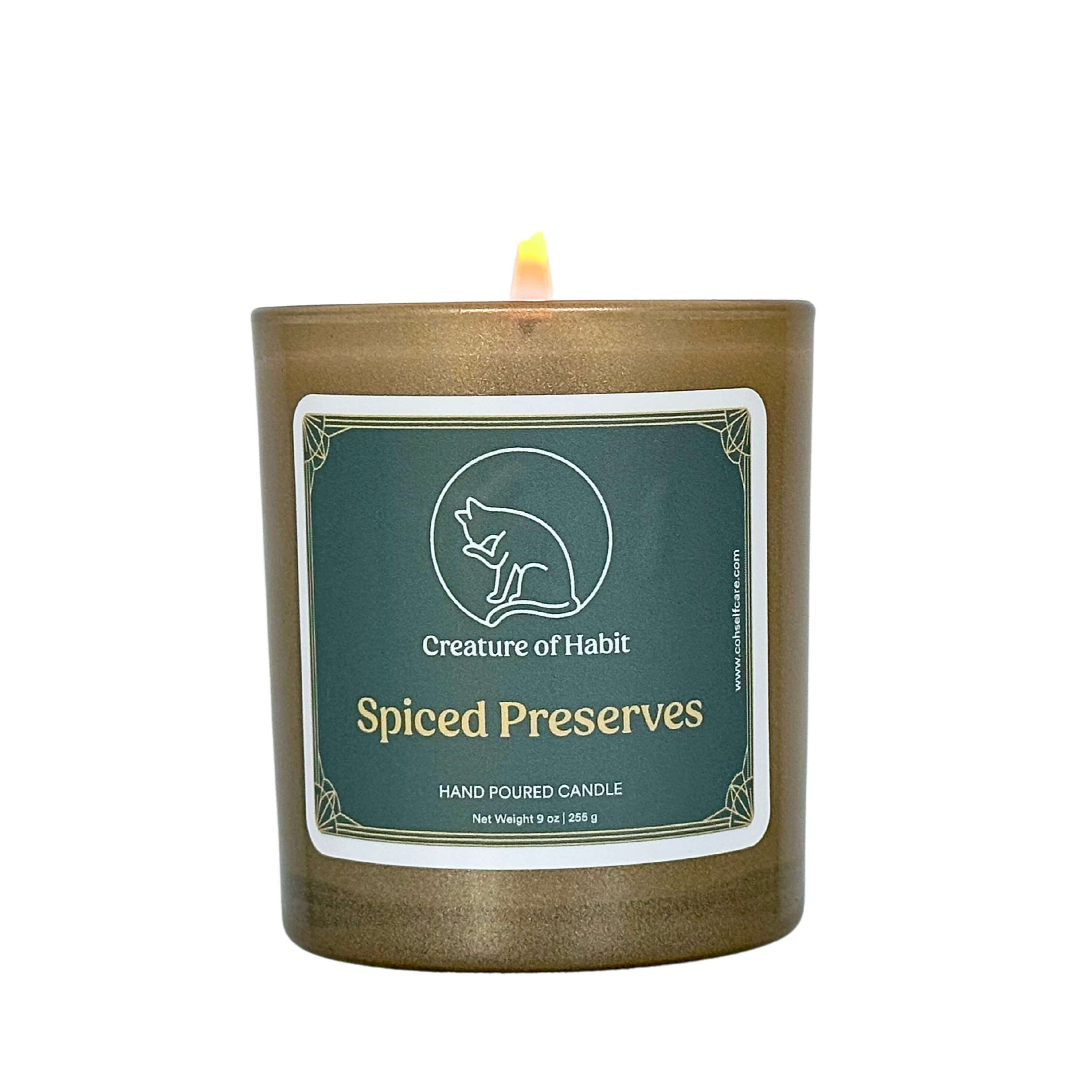 A lit soy candle within a golden vessel is against a white background. The label is greyish cyan featuring the logo of a white cat silhouette, the name of the company Creature of Habit, and the scent name Spiced Preserves.