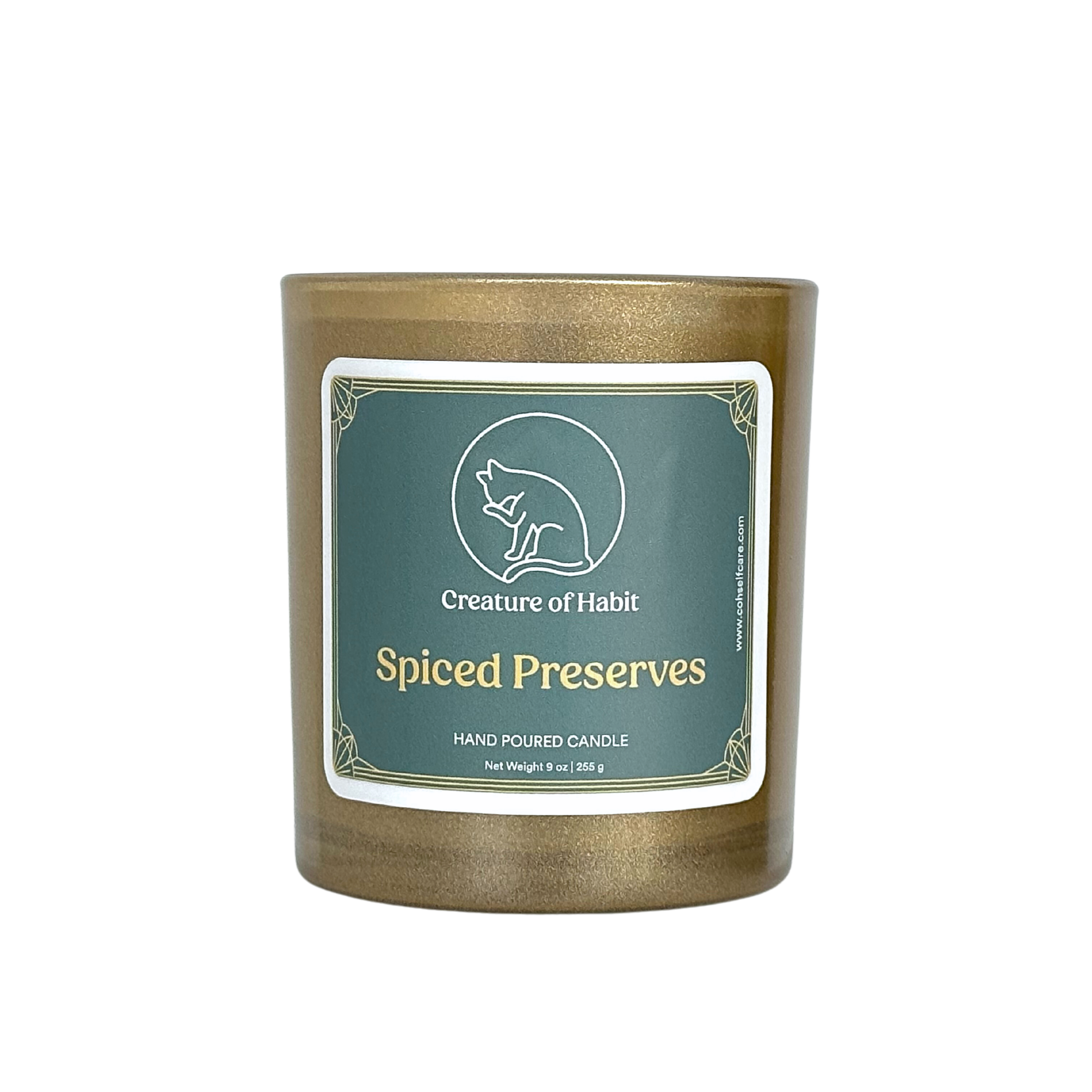 An unlit soy candle within a golden vessel is against a white background. The label is a greyish cyan featuring the logo of a white cat silhouette, the name of the company Creature of Habit, and the scent name Spiced Preserves.
