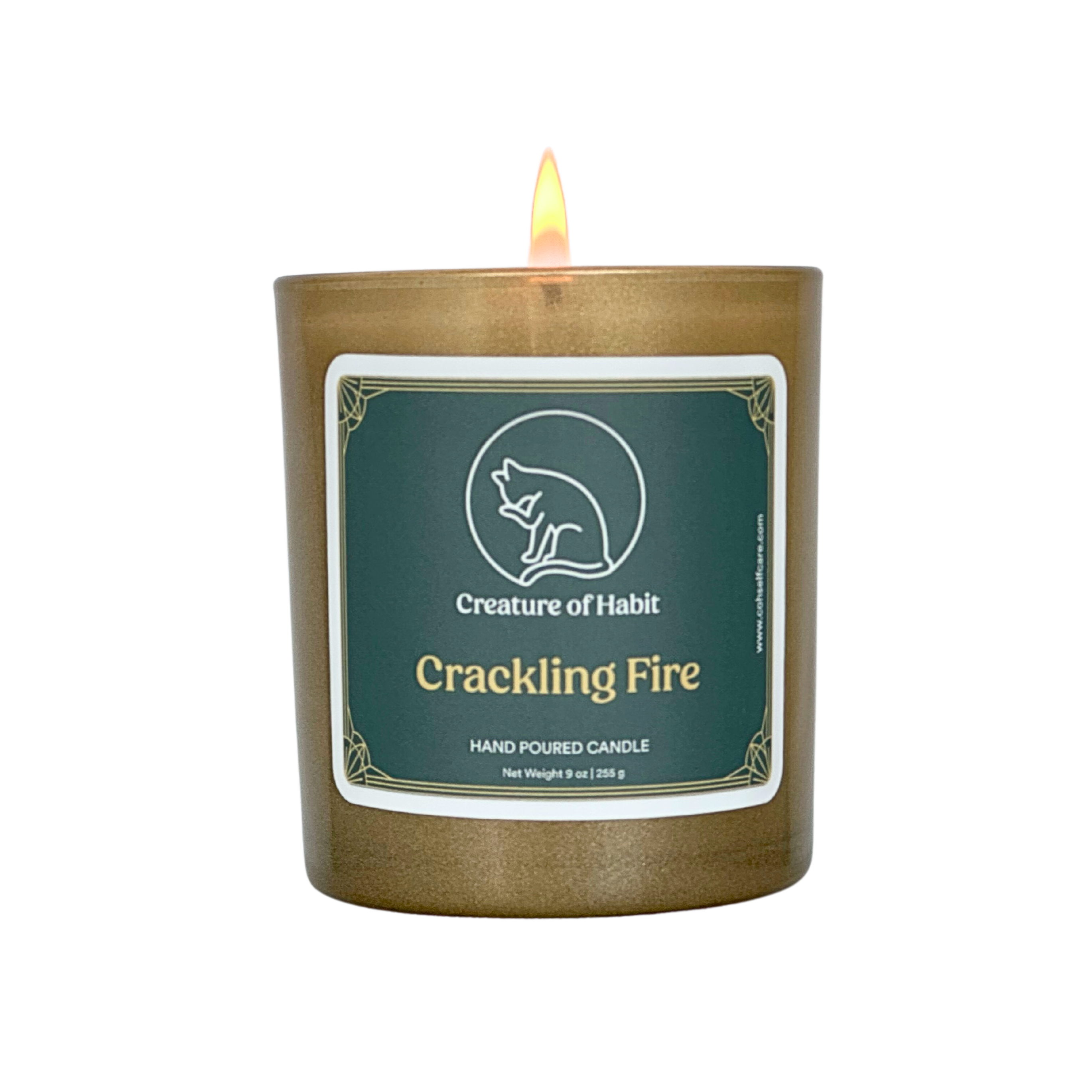 A lit soy candle within a golden vessel is against a white background. The label is greyish cyan featuring the logo of a white cat silhouette, the name of the company Creature of Habit, and the scent name Crackling Fire.