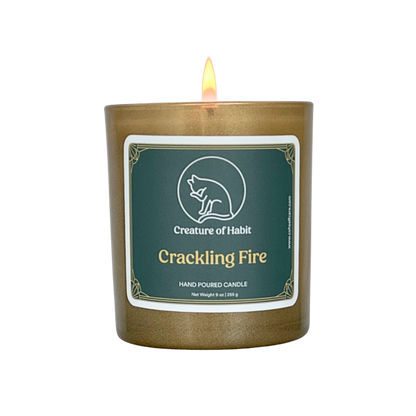 A lit soy candle within a golden vessel is against a white background. The label is greyish cyan featuring the logo of a white cat silhouette, the name of the company Creature of Habit, and the scent name Crackling Fire.