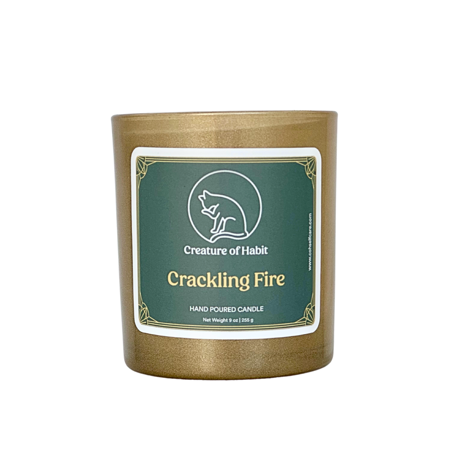 An unlit soy candle within a golden vessel is against a white background. The label is a greyish cyan featuring the logo of a white cat silhouette, the name of the company Creature of Habit, and the scent name Crackling Fire.