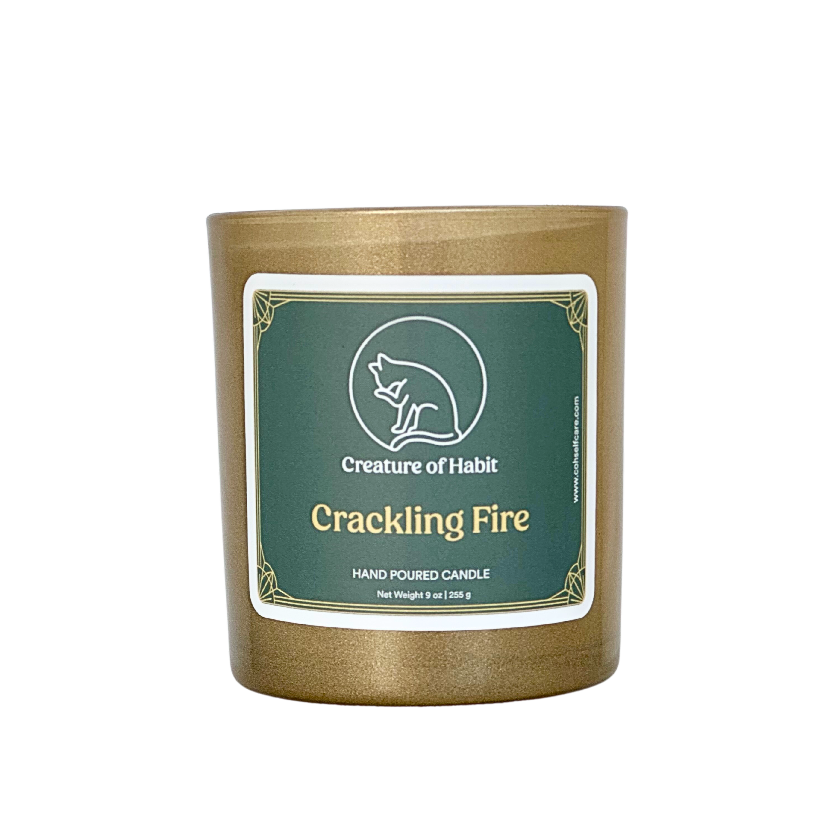 An unlit soy candle within a golden vessel is against a white background. The label is a greyish cyan featuring the logo of a white cat silhouette, the name of the company Creature of Habit, and the scent name Crackling Fire.