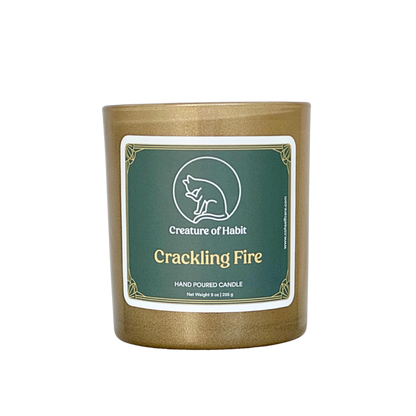 An unlit soy candle within a golden vessel is against a white background. The label is a greyish cyan featuring the logo of a white cat silhouette, the name of the company Creature of Habit, and the scent name Crackling Fire.