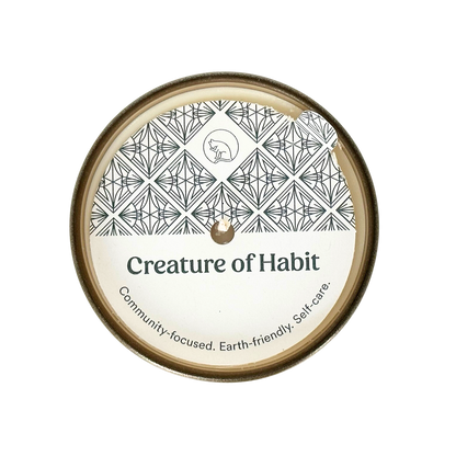 A top-down view of a candle is shown with a dust cover. The dust cover features the Creature of Habit logo, a silhouette of a cat, within a repeating art deco pattern. The company name Creature of Habit is featured along with the tagline, &quot;Community-focused. Earth-friendly. Self-care.”