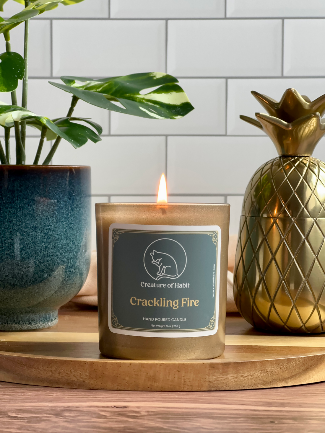 A lit soy candle within a golden vessel is placed on top of a wooden tray, next to a small houseplant and golden pineapple paperweight. The label is greyish cyan featuring the logo of a white cat silhouette, the name of the company Creature of Habit, and the scent name Crackling Fire.