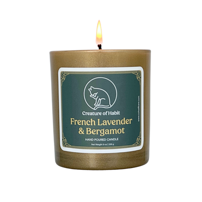 A lit soy candle within a golden vessel is against a white background. The label is greyish cyan featuring the logo of a white cat silhouette, the name of the company Creature of Habit, and the scent name French Lavender &amp; Bergamot.