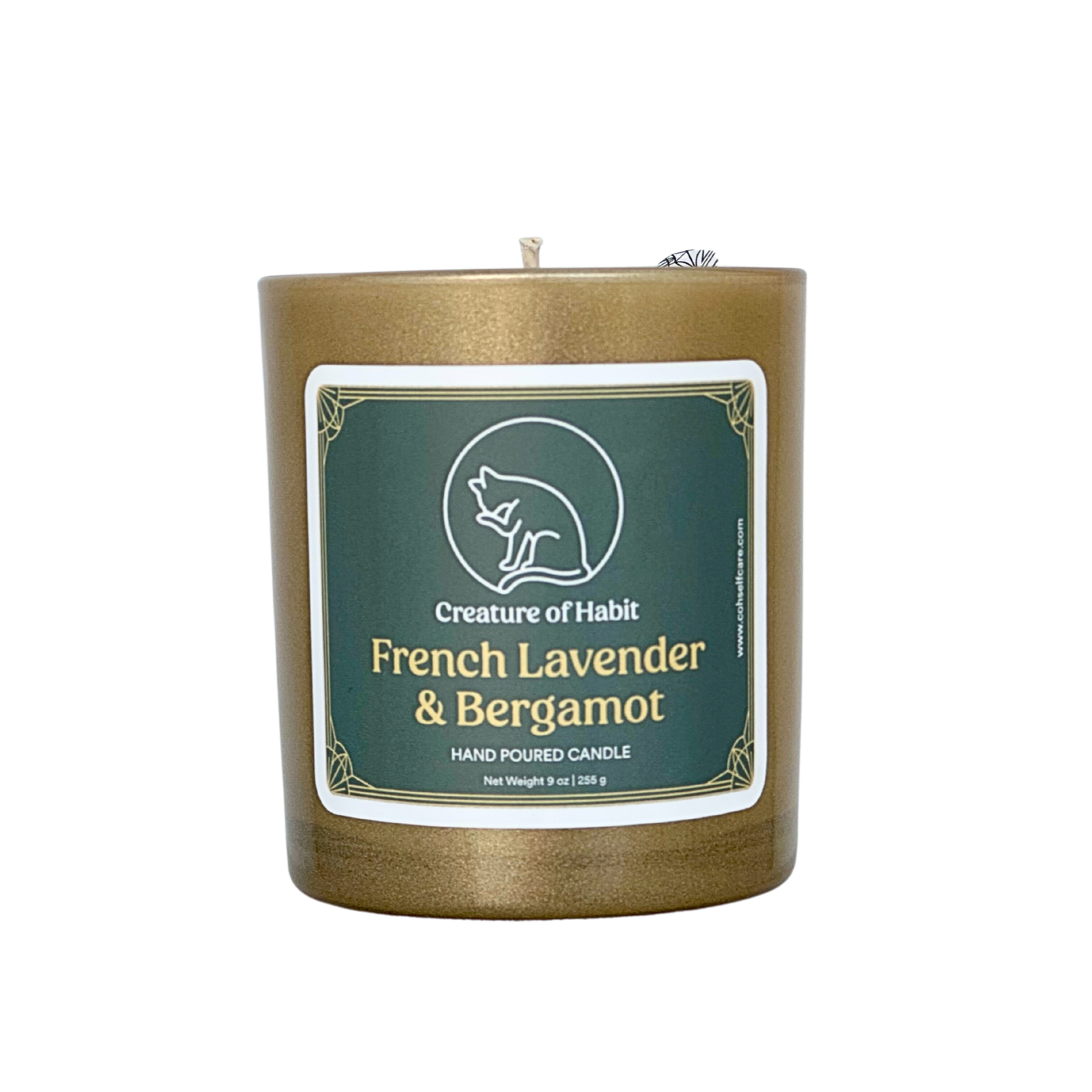 An unlit soy candle within a golden vessel is against a white background. The label is a greyish cyan featuring the logo of a white cat silhouette, the name of the company Creature of Habit, and the scent name French Lavender &amp; Bergamot.