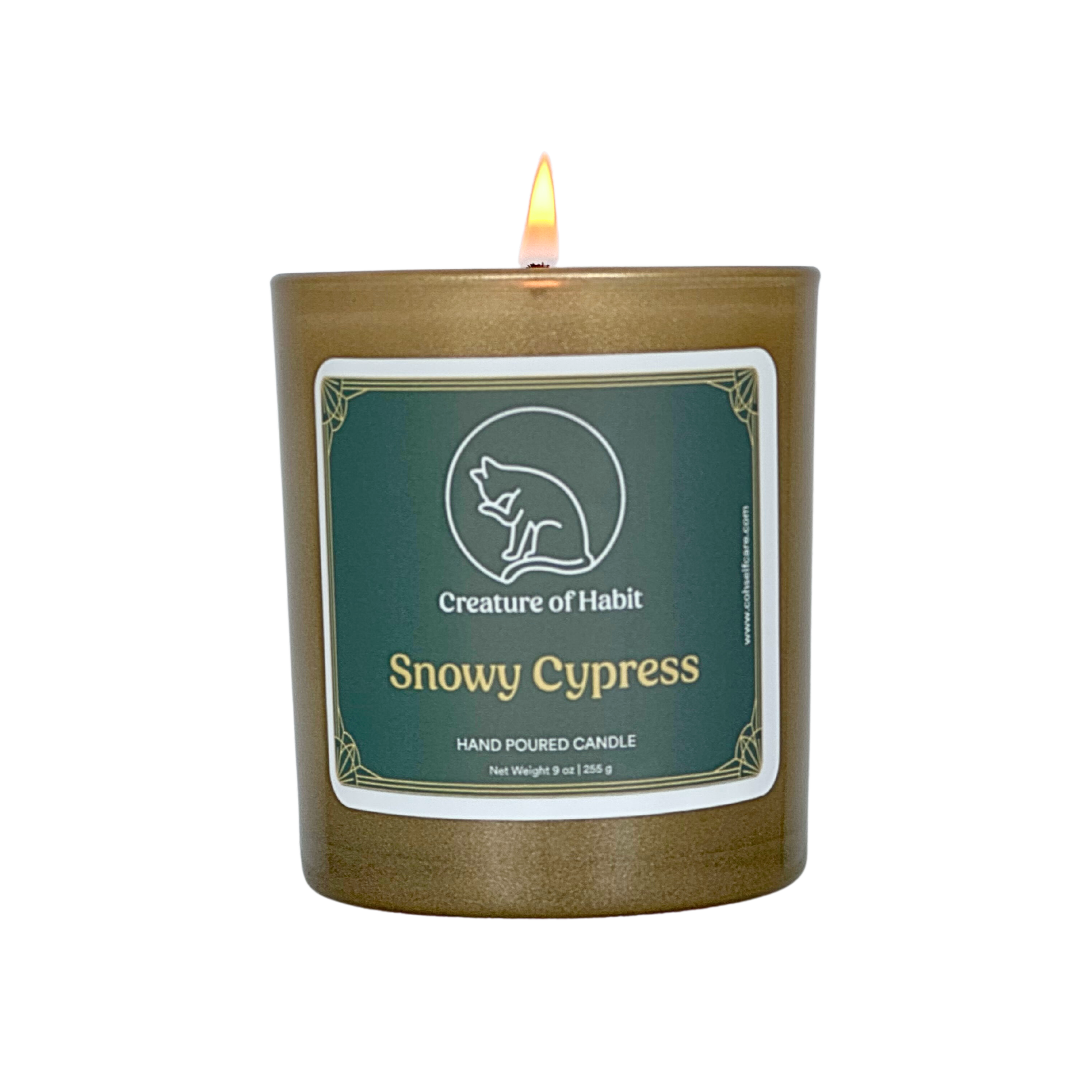 A lit soy candle within a golden vessel is against a white background. The label is greyish cyan featuring the logo of a white cat silhouette, the name of the company Creature of Habit, and the scent name Snowy Cypress.