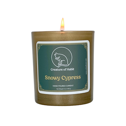 A lit soy candle within a golden vessel is against a white background. The label is greyish cyan featuring the logo of a white cat silhouette, the name of the company Creature of Habit, and the scent name Snowy Cypress.