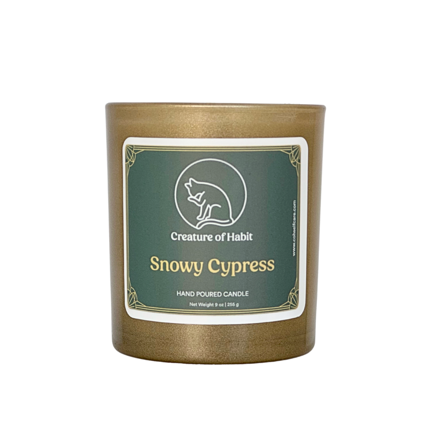 An unlit soy candle within a golden vessel is against a white background. The label is a greyish cyan featuring the logo of a white cat silhouette, the name of the company Creature of Habit, and the scent name Snowy Cypress.