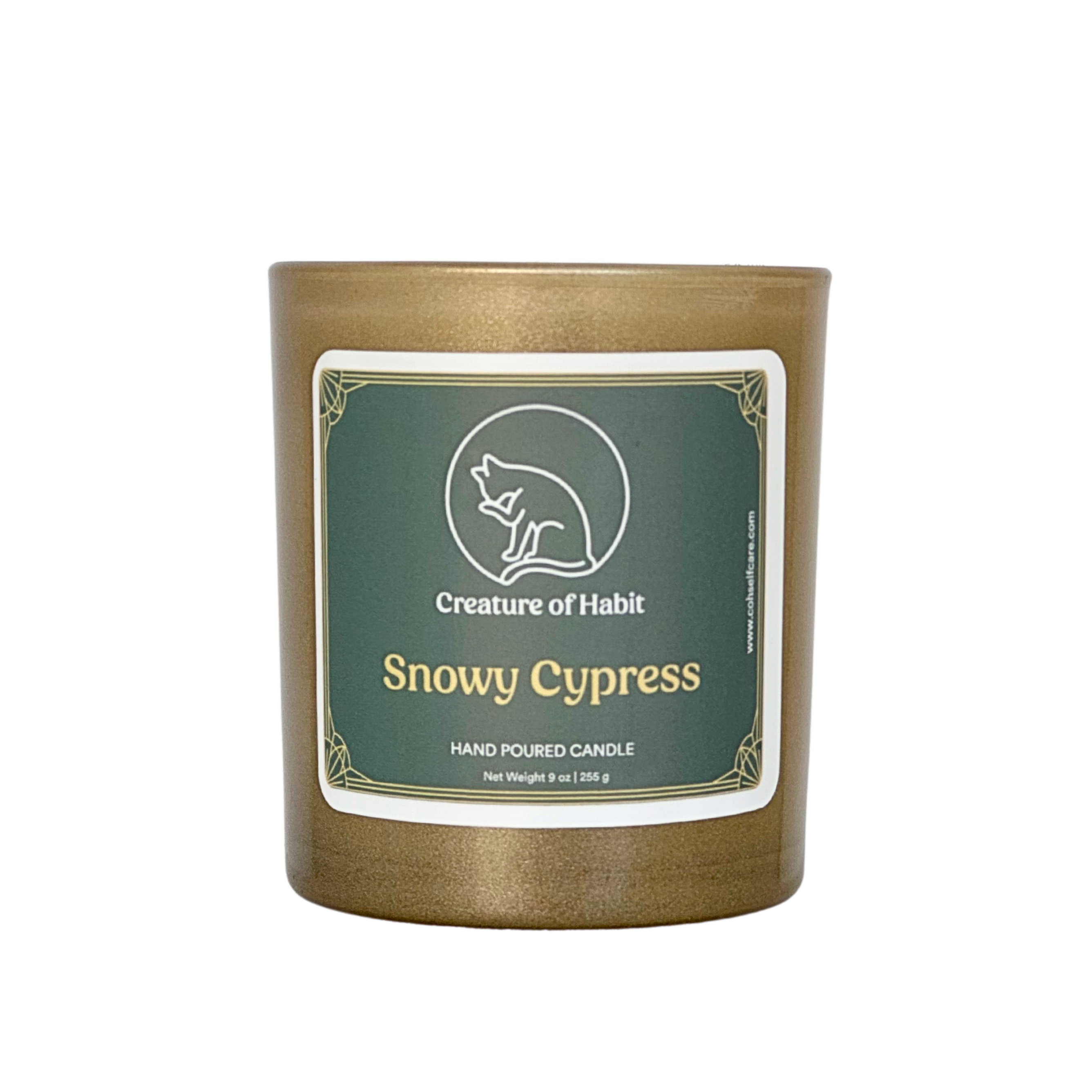 An unlit soy candle within a golden vessel is against a white background. The label is a greyish cyan featuring the logo of a white cat silhouette, the name of the company Creature of Habit, and the scent name Snowy Cypress.