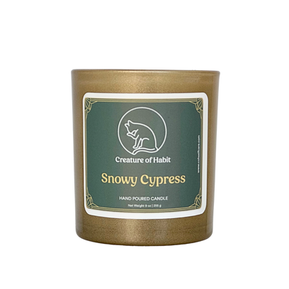 An unlit soy candle within a golden vessel is against a white background. The label is a greyish cyan featuring the logo of a white cat silhouette, the name of the company Creature of Habit, and the scent name Snowy Cypress.