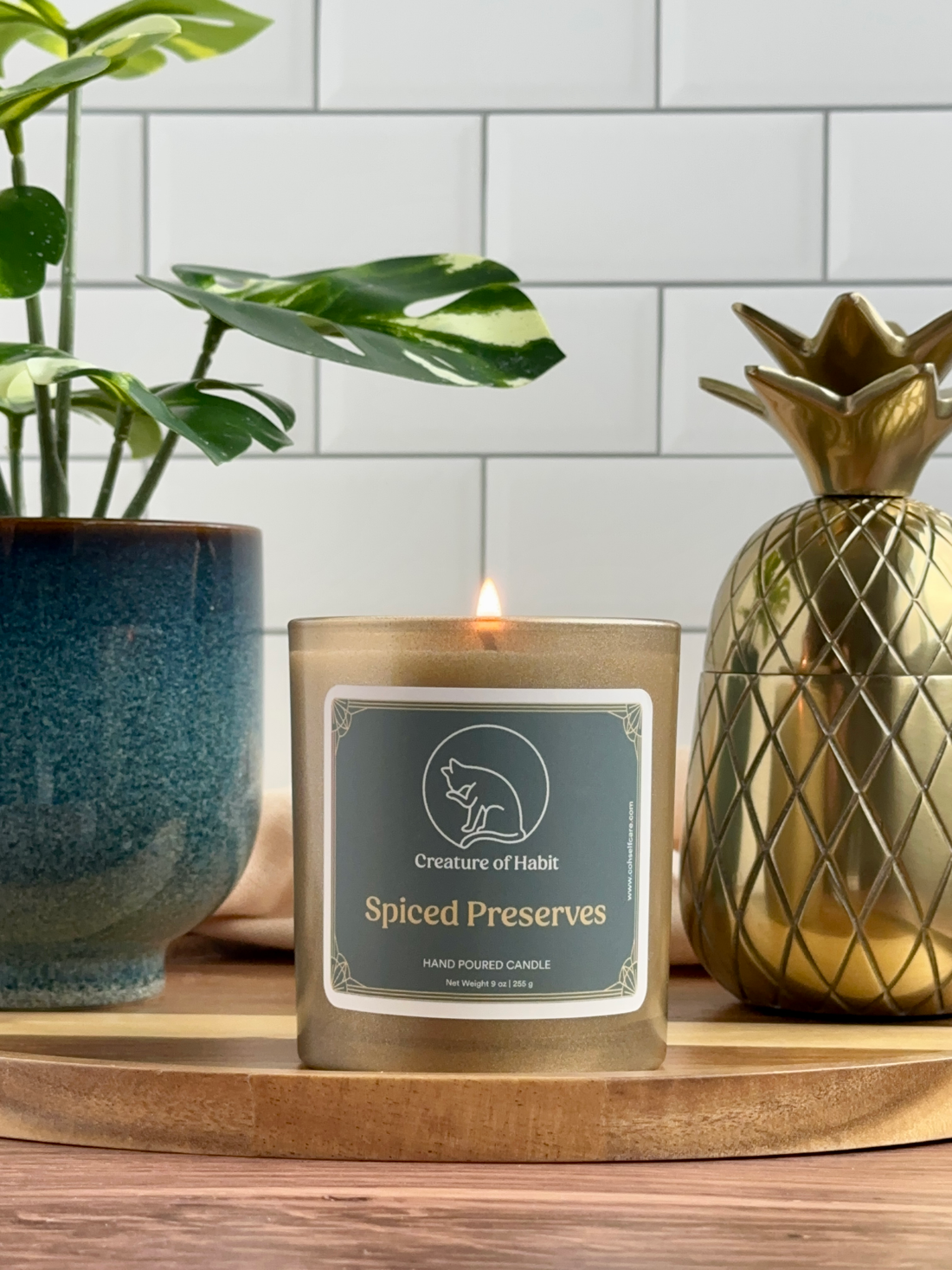 A lit soy candle within a golden vessel is placed on top of a wooden tray, next to a small houseplant and golden pineapple paperweight. The label is greyish cyan featuring the logo of a white cat silhouette, the name of the company Creature of Habit, and the scent name Spiced Preserves.